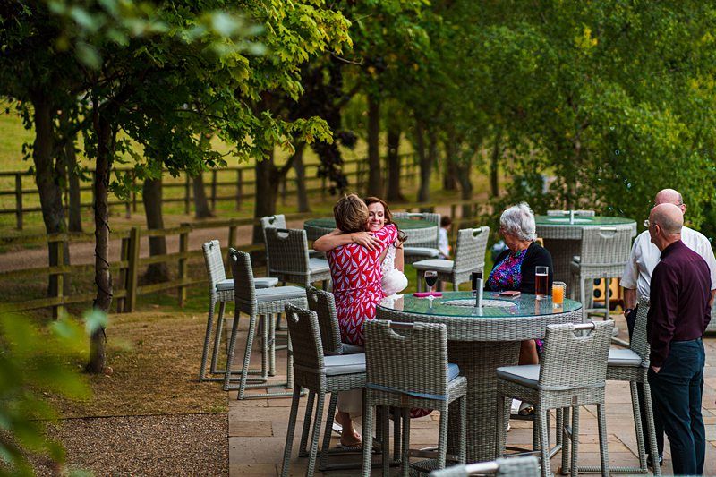 A group of people sits and interacts around wicker patio tables and chairs in an outdoor setting surrounded by trees and a wooden fence. One person wearing a pink dress hugs another person, while the others engage in conversation. Drinks are visible on the tables. Image by Documentary Wedding Photographer Lewis Bishop Photography.