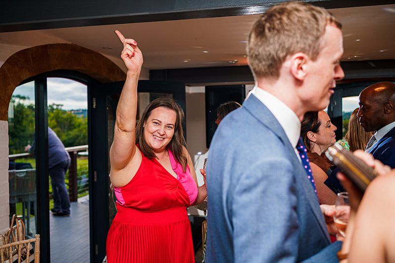 A woman in a red dress stands indoors with one arm raised, smiling at the camera. She is among a group of people socializing. In the foreground, a man in a suit is visible from the back, engaging in conversation. The background shows large windows and outside greenery. Image by Documentary Wedding Photographer Lewis Bishop Photography.