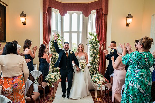 A newlywed couple walks down the aisle, smiling and waving, in an elegantly decorated room with flowers. Guests on either side cheer and clap. The bride wears a white gown, and the groom is in a dark suit. Large windows and red curtains frame the background. Image by Documentary Wedding Photographer Lewis Bishop Photography.