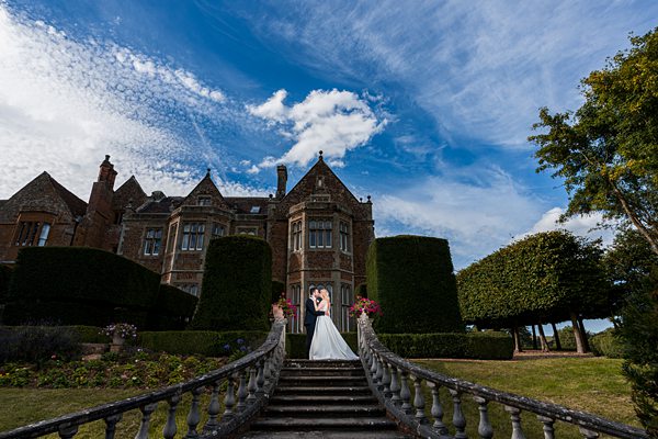 Couple embracing in front of historic manor under blue sky.