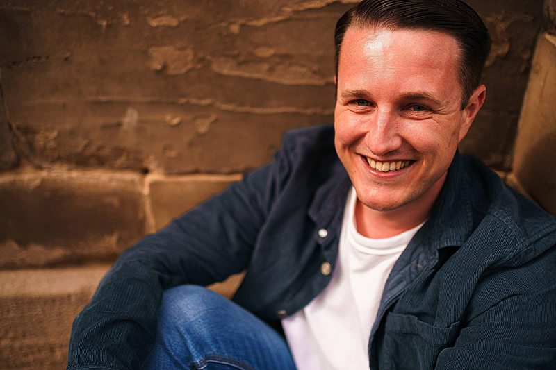 Smiling man sitting casually against a brick wall.