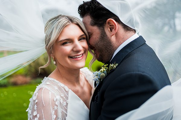Bride and groom smiling under veiled embrace.