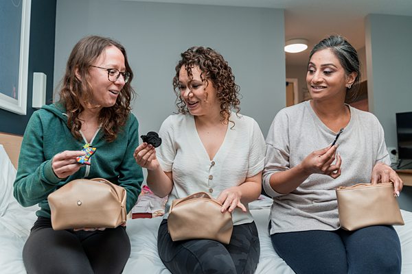 Three women laughing and sharing snacks on bed.