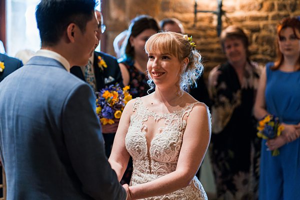 A bride and groom smiling at each other during their wedding ceremony. The bride is wearing a intricately designed lace wedding dress and holding the groom's hands. The groom is in a light blue suit. They are surrounded by friends and family, some holding flowers. Image by Documentary Wedding Photographer Lewis Bishop Photography.