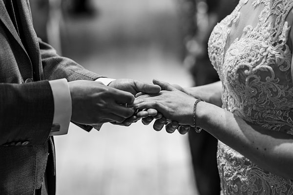 A black-and-white photo showing a close-up of a bride and groom holding hands during a wedding ceremony. The groom is placing a ring on the bride's finger. Both are dressed in formal attire, with the bride wearing a lace wedding dress. Image by Documentary Wedding Photographer Lewis Bishop Photography.