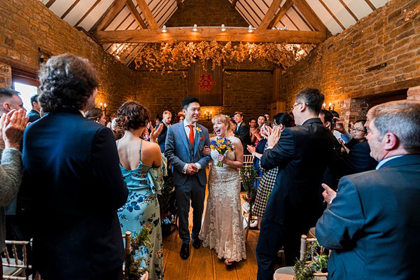 A joyful bride and groom walk down the aisle in a rustic, warmly lit venue adorned with wooden beams and floral arrangements. Guests on either side stand and applaud, celebrating the couple's union. The bride holds a bouquet, and both are smiling brightly. Image by Documentary Wedding Photographer Lewis Bishop Photography.