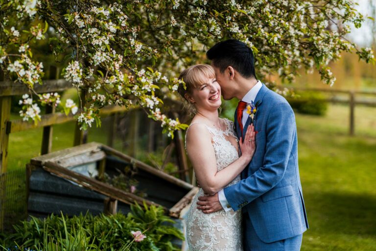 A joyful couple poses under a blooming tree. The bride, in an ornate white dress, smiles brightly as the groom, in a light blue suit, kisses her cheek. They stand in a lush garden with wooden structures and greenery in the background. Image by Documentary Wedding Photographer Lewis Bishop Photography.