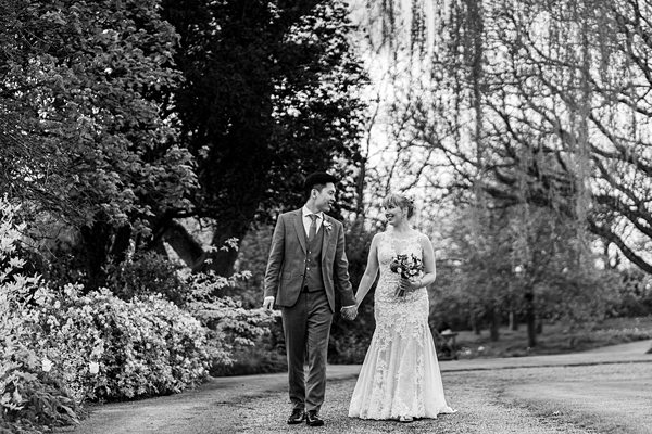A bride in a lace wedding dress and a groom in a suit walk hand in hand through a garden. They are smiling at each other, surrounded by blossoming trees and greenery. The photo is in black and white, creating a timeless and romantic atmosphere. Image by Documentary Wedding Photographer Lewis Bishop Photography.