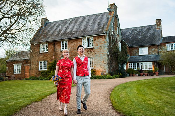 A couple dressed in red traditional attire walks hand in hand on a gravel path leading to a large stone house with a garden. The woman carries a bouquet of flowers, and both are smiling, with green lawns on either side of the path and a large tree to the left. Image by Documentary Wedding Photographer Lewis Bishop Photography.
