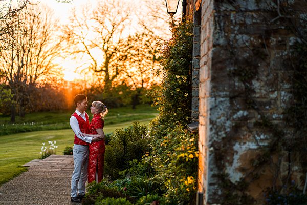 A couple, dressed in traditional Chinese attire, stands next to a stone wall adorned with ivy and foliage. The man is in a red vest and white shirt, while the woman wears a red dress. They embrace as the sun sets, casting a warm glow over the lush garden scenery. Image by Documentary Wedding Photographer Lewis Bishop Photography.