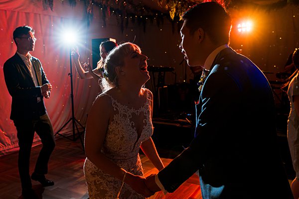 A couple in formal attire, with the bride in a white lace dress and the groom in a suit, are laughing and holding hands while dancing. The scene is illuminated by colorful, dramatic lighting with guests and photographers in the background. Image by Documentary Wedding Photographer Lewis Bishop Photography.