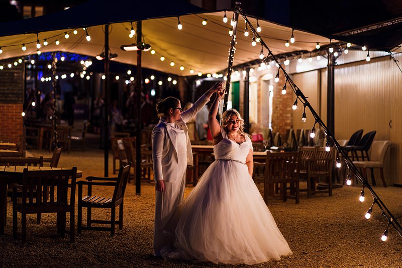 Bride and friend dancing under fairy lights at evening wedding.