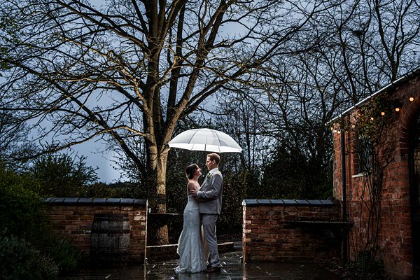 A newlywed couple stands close under a glowing umbrella on a rainy evening. They are outside near a brick building adorned with string lights. The background features leafless trees and a moody, overcast sky. The bride wears a white dress and the groom a light grey suit. Image by Documentary Wedding Photographer Lewis Bishop Photography.