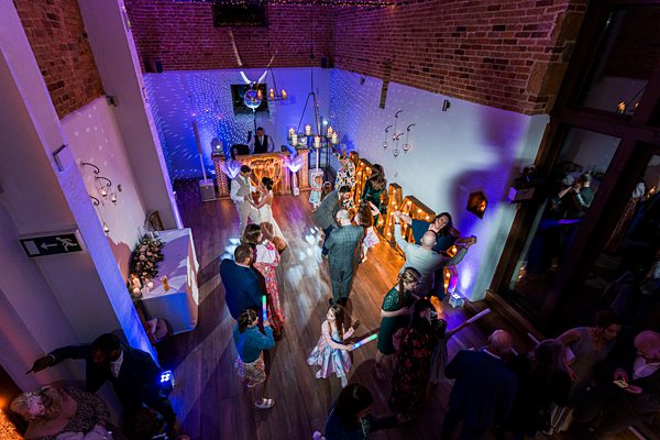 A lively wedding reception with guests dancing and mingling in a decorated hall with brick walls. Fairy lights and candles add to the ambiance. A DJ booth is set up on one end, and a bride and groom can be seen dancing together in the center of the room. Image by Documentary Wedding Photographer Lewis Bishop Photography.