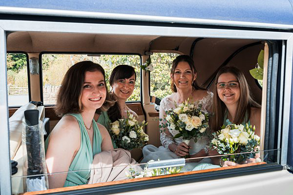 Bridesmaids and bride smiling in vintage car with flowers.