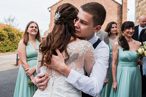 Bride and groom embrace at wedding, guests smiling.