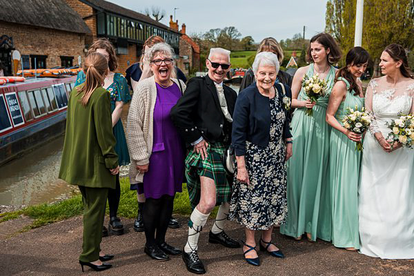 Wedding guests smiling by canal, man in kilt.