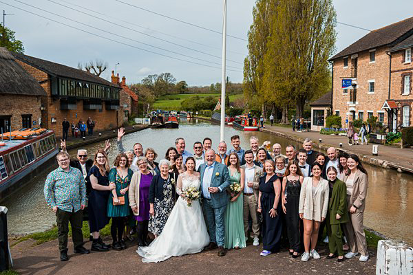 Wedding party by canal with boat and guests smiling.