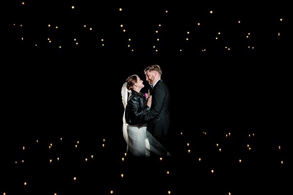 A bride and groom share an intimate moment under a canopy of small, warmly lit string lights. The bride wears a white dress and veil, paired with a black leather jacket, while the groom is in a dark suit. The background is dark, making the lights shine brightly. Image by Documentary Wedding Photographer Lewis Bishop Photography.