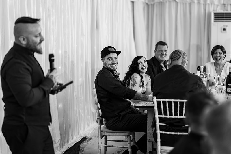 Groom smiling at wedding speech, guests laugh, event celebration.