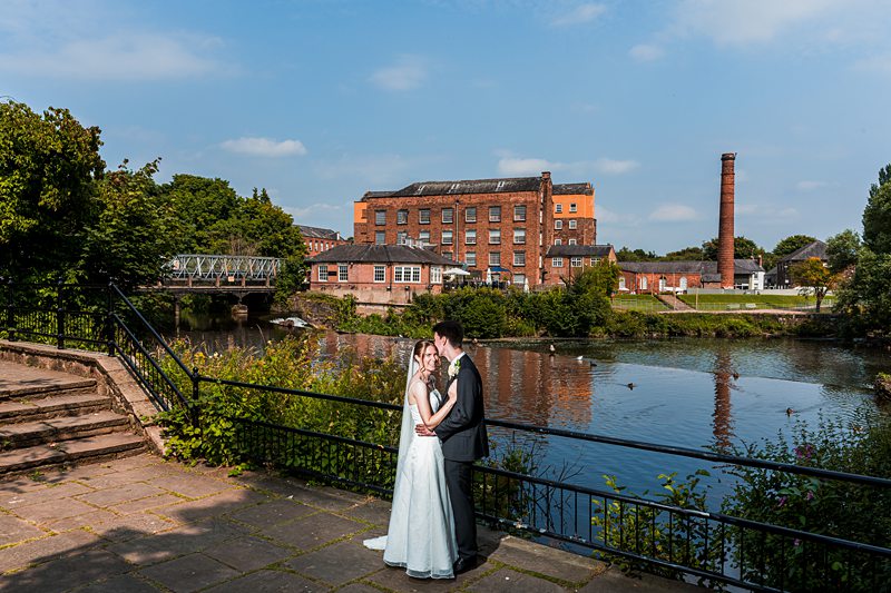 Newlyweds by river with historic mill backdrop.