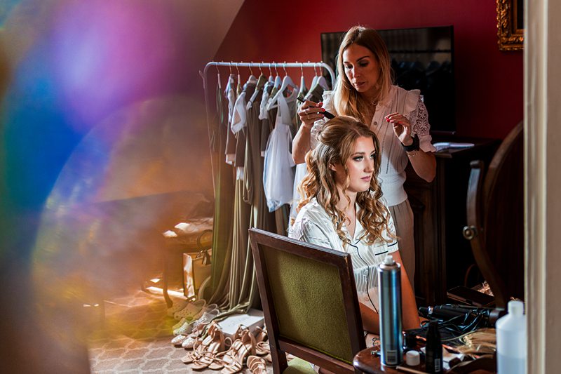 Bridesmaid getting ready with hair stylist, dresses in background.