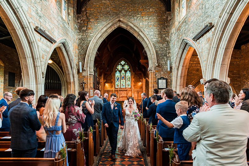 Wedding couple walking down church aisle, guests clapping.
