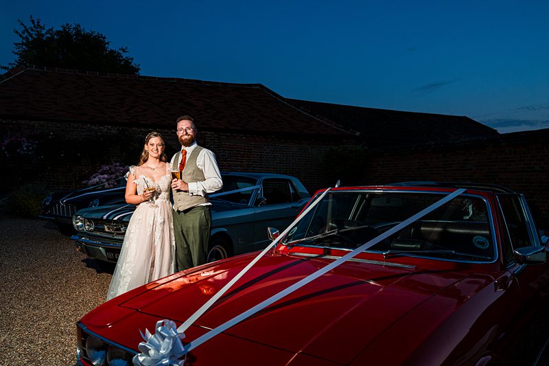 Newlyweds with champagne beside decorated vintage cars.