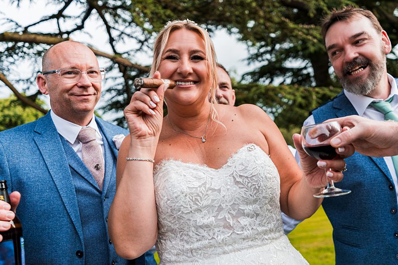 Bride celebrating with cigar and guests outdoors.