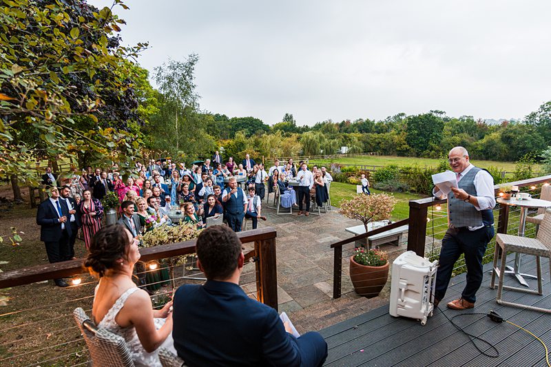 Wedding speech in countryside setting with guests listening.