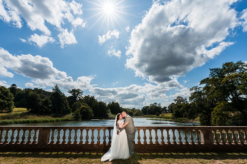 Wedding couple embracing by riverside under sunny sky.
