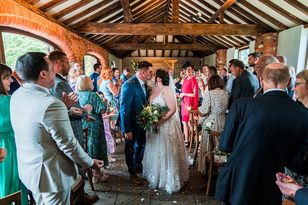 Bride and groom kiss, guests applaud in rustic venue.