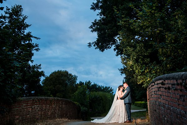 Newlyweds embrace on stone bridge at dusk.
