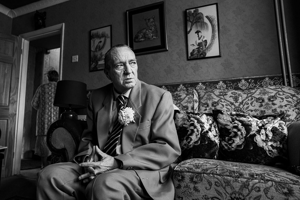 Elderly man in suit, seated on floral sofa.