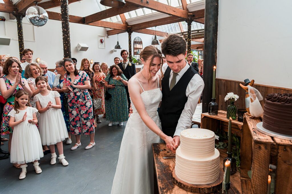 Couple cutting wedding cake with guests clapping.