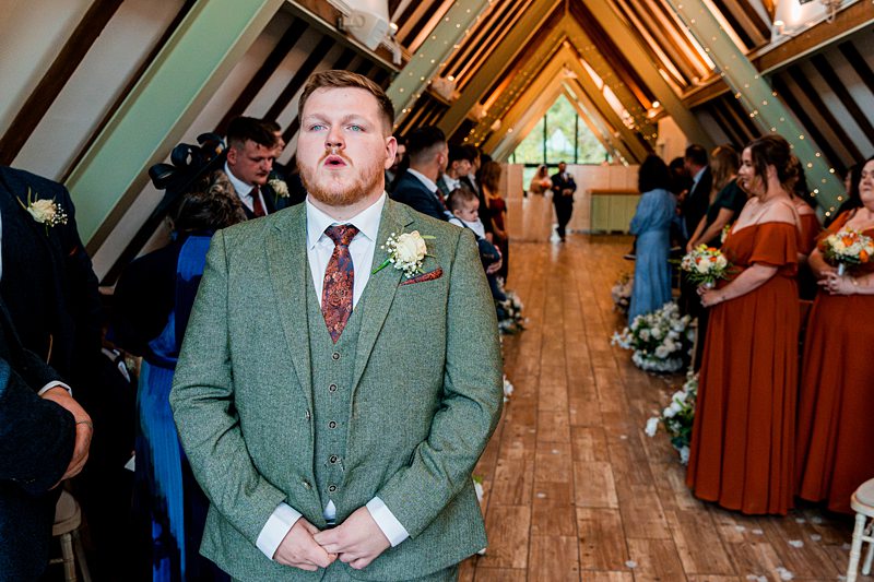 Groom in green suit awaits bride at aisle.