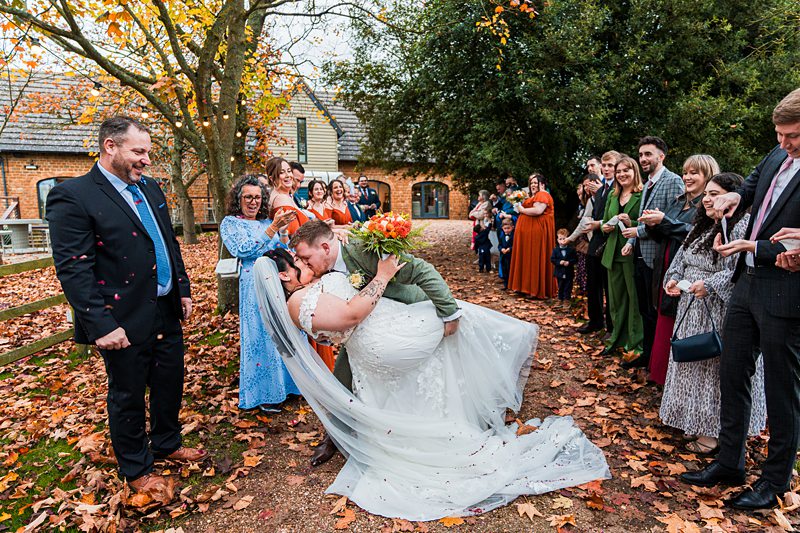 Bride and groom kissing, surrounded by guests and foliage.