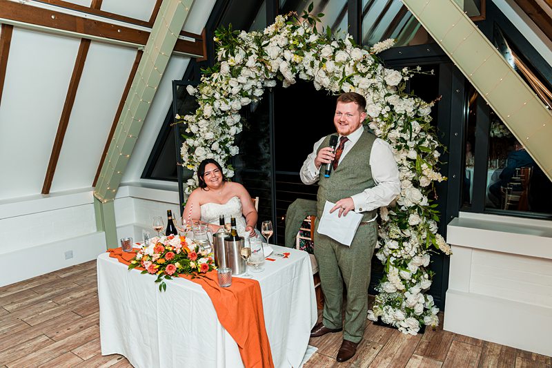 Bride and groom delivering wedding speech, floral arch backdrop.