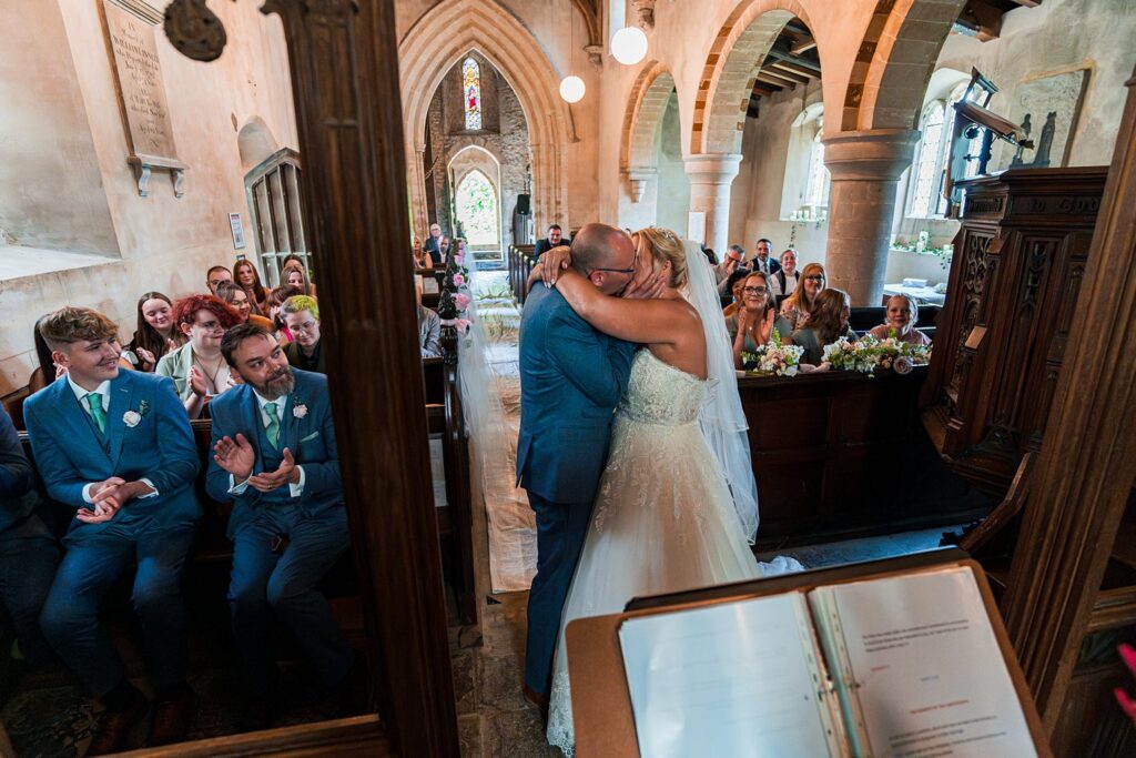 Newlyweds kissing in a church, guests clapping.