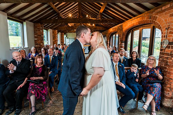 Couple kissing at indoor wedding ceremony, guests clapping.