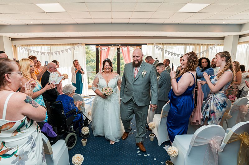 Newlyweds walking down aisle, guests applauding.