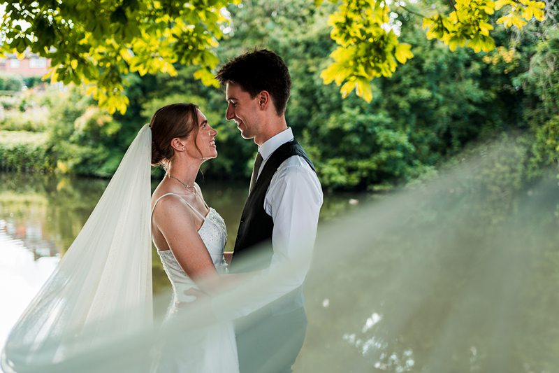 Bride and groom smiling by a leafy river.