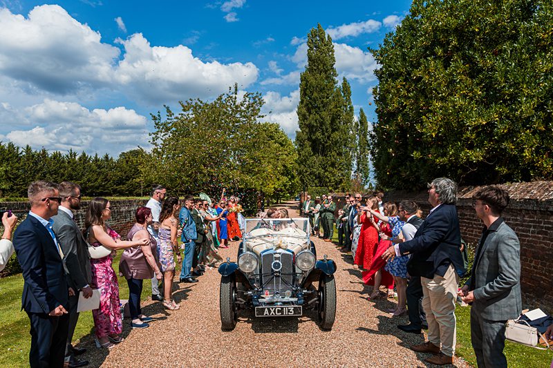 Wedding guests cheer vintage car departure.