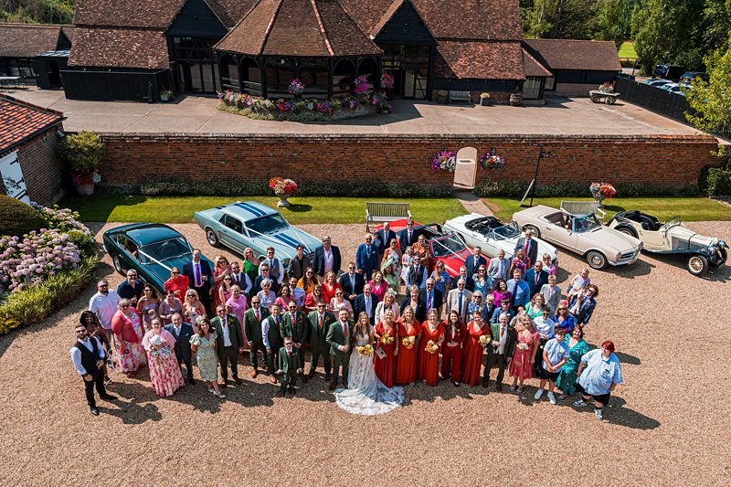 Wedding guests pose outside vintage cars and barn.