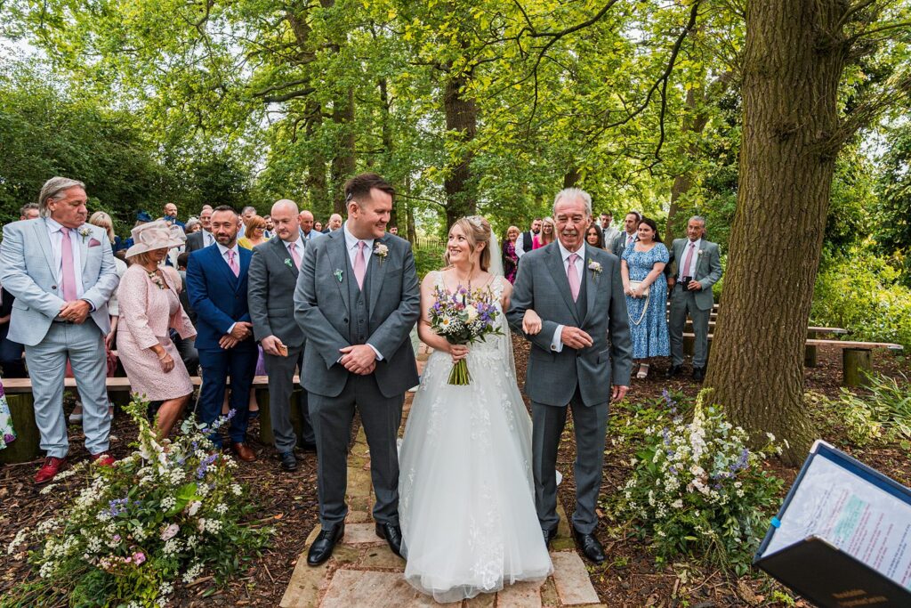 Bride walking down forest aisle with guests watching.