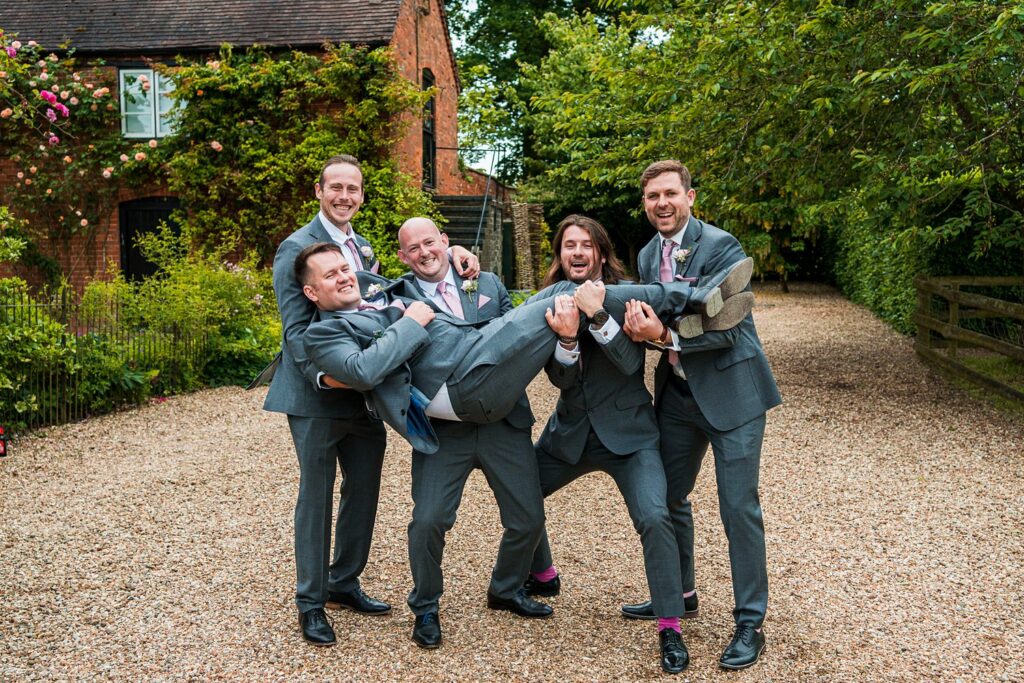Groomsmen in suits lifting friend outdoors, smiling.