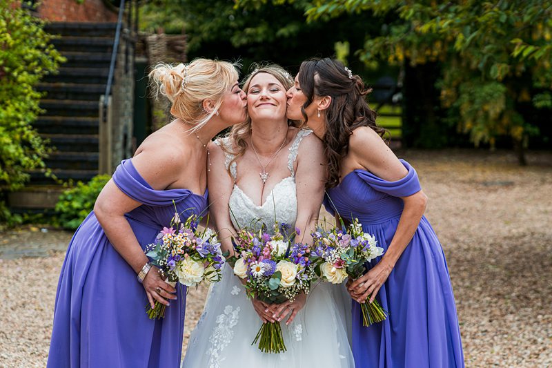 Bride with bridesmaids kissing her cheeks, smiling.