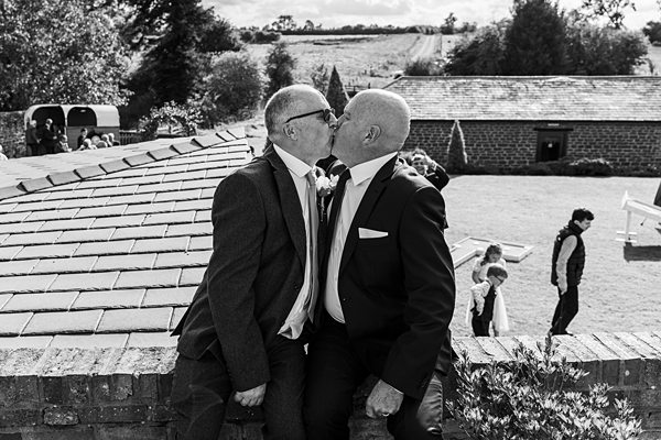 Two men in suits kissing at outdoor event.