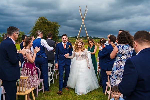 Bride and groom walk down outdoor aisle, cloudy sky.