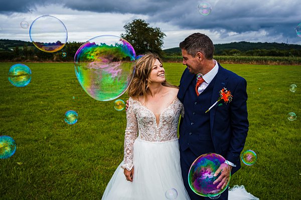 Bride and groom smiling surrounded by bubbles outdoors.